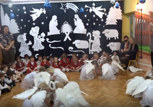 Na tle bożonarodzeniowej dekoracji w czteroosobowych zespołach dzieci w strojach aniołów tańczą kucając.